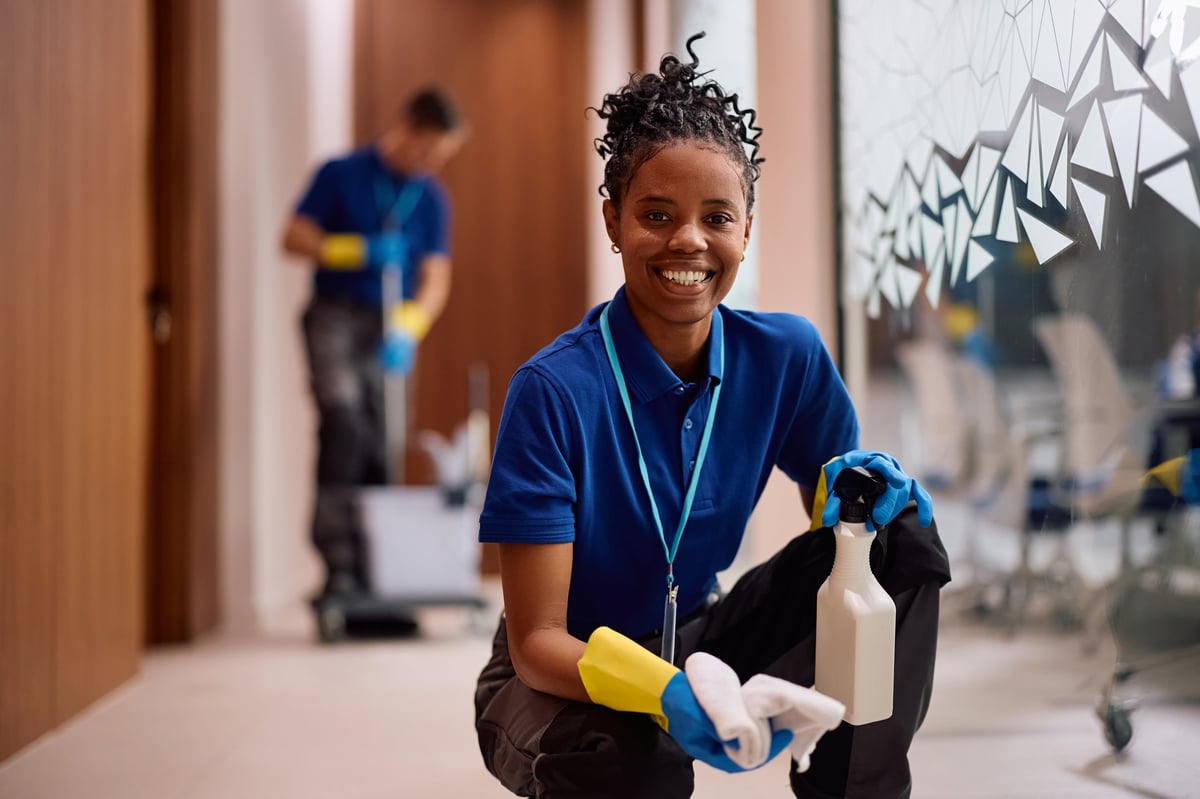 Happy African American female cleaner wiping glass walls in hallway of an office building